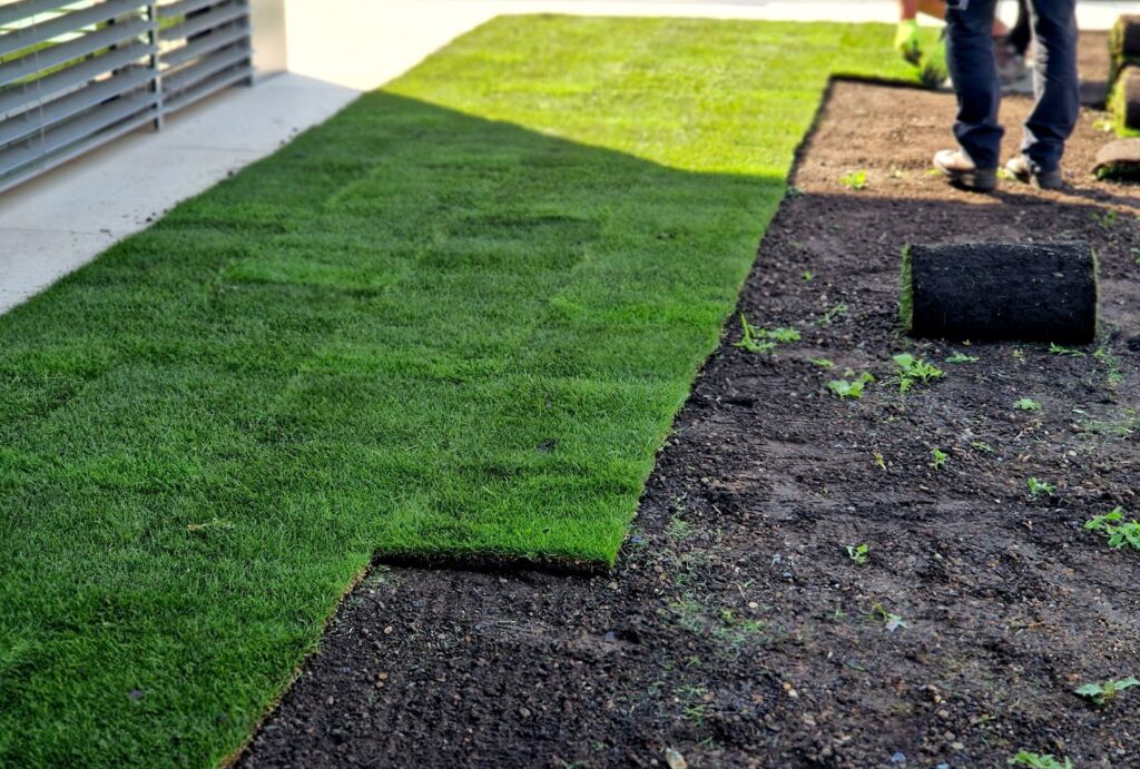 Person installing rolled sod next to a prepared soil area near a building.