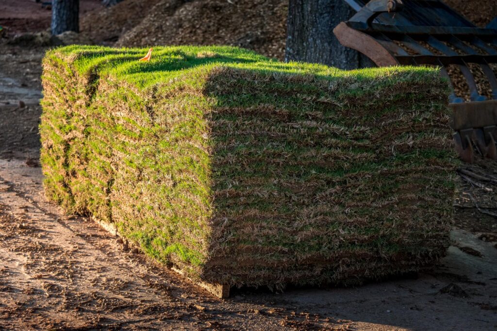 A large, rectangular pallet stacked high with rolls of fresh, green sod sits on a patch of dirt and gravel in a partially shaded outdoor area.