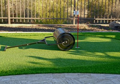 A sod roller on an artificial turf backyard putting green. A green artificial turf putting green with a hole, a red flag on a white pole, and a black lawn roller lying on its side.